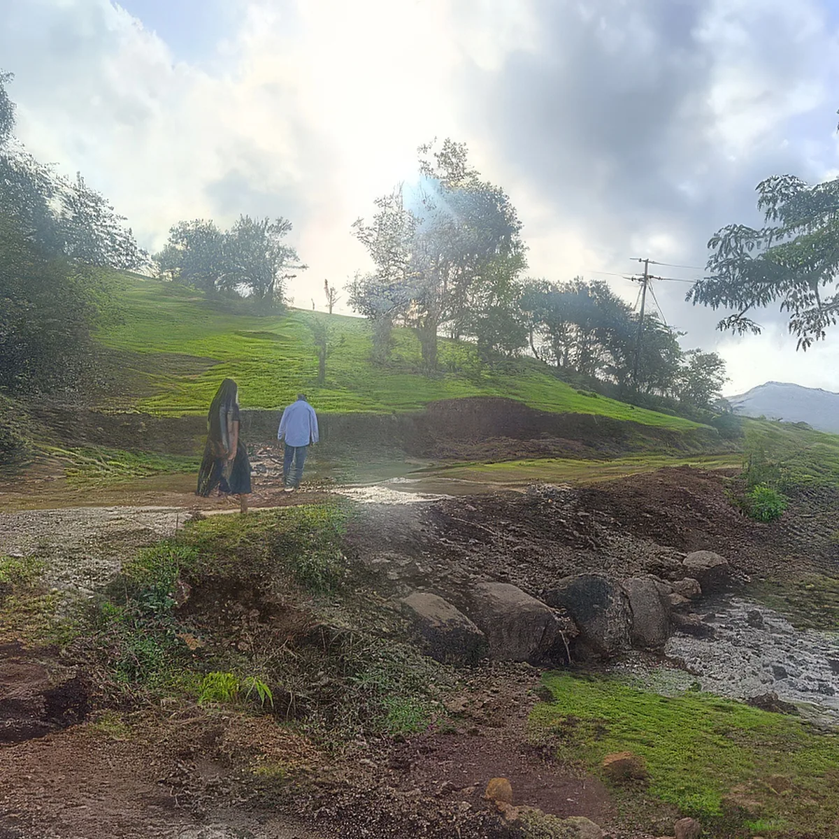 Two people crossing a stream on a dirt path, with a utility pole beyond.